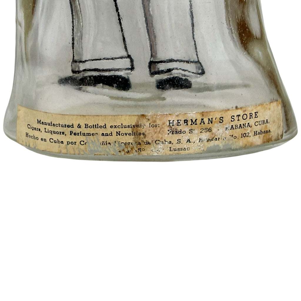 Close-up of the lower portion of a vintage Cuban glass bottle with a beige paper label reading 'HERMAN'S STORE' and manufacturing details for Habana, Cuba.