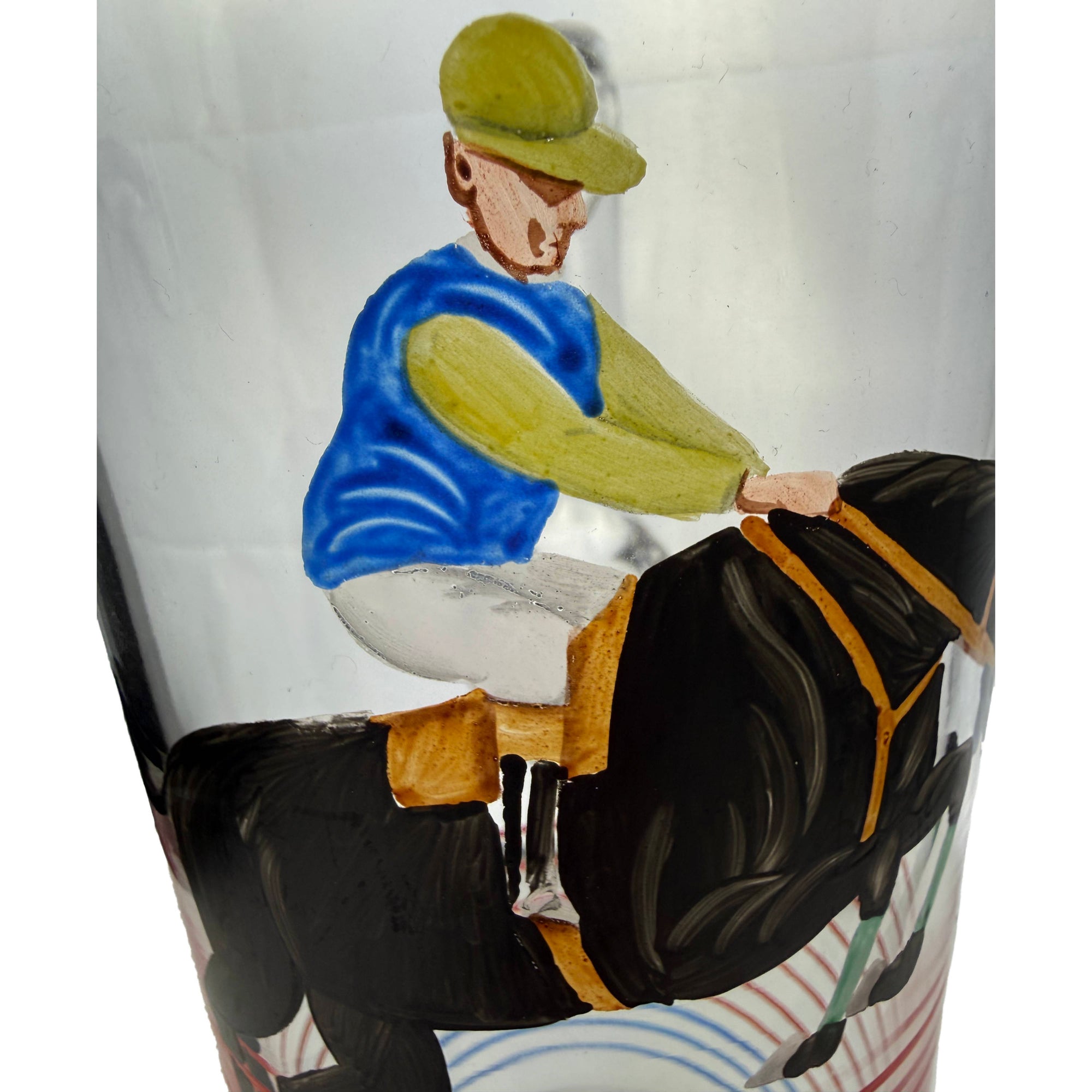 A close-up of a glass martini pitcher featuring a painted illustration of a jockey riding a horse. The artwork depicts the jockey in a blue vest and green cap, and the horse in black with gold accents