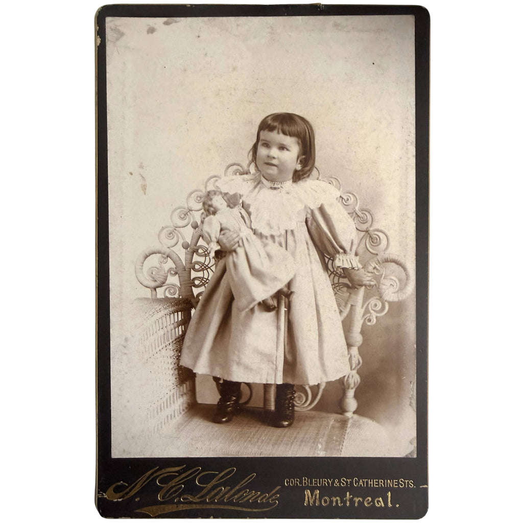 An antique sepia-toned portrait photograph featuring a young girl in a ruffled dress holding a doll
