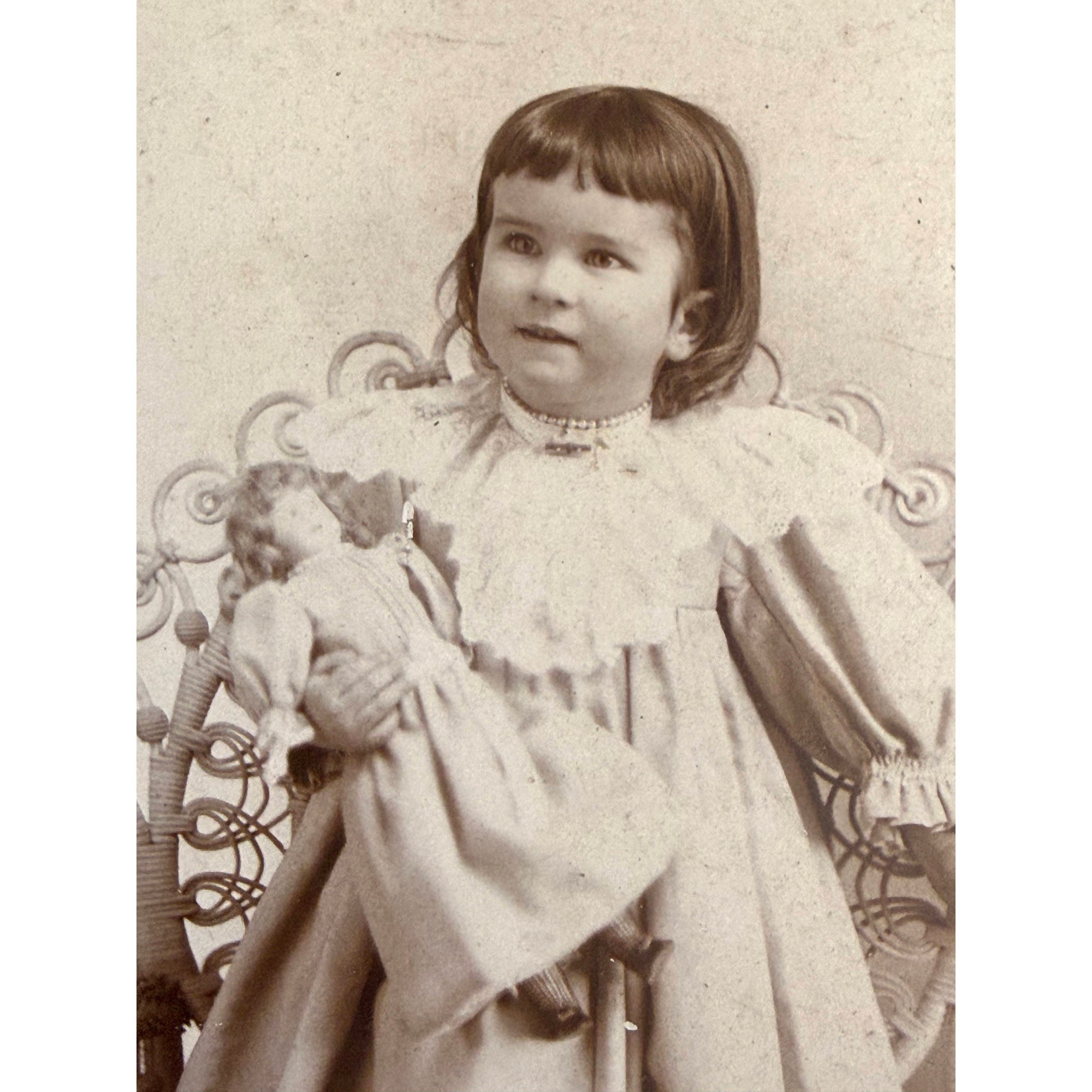Vintage sepia-toned portrait of a young girl wearing a high-neck ruffled dress and holding a doll, in front of an ornate chair.
