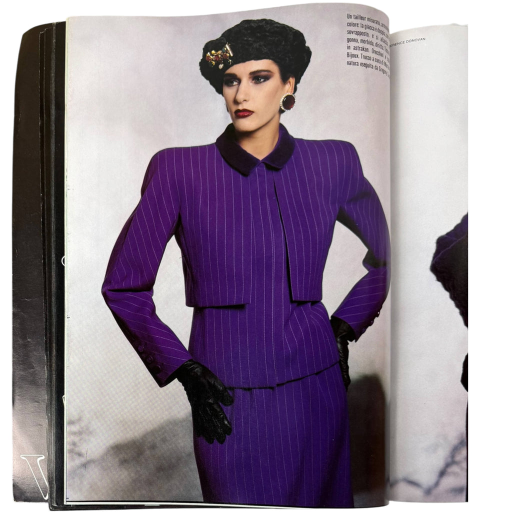 An open fashion magazineshowing a model in a Valentino purple pinstripe suit with a black collar, black hat, and black gloves on a light gray page.