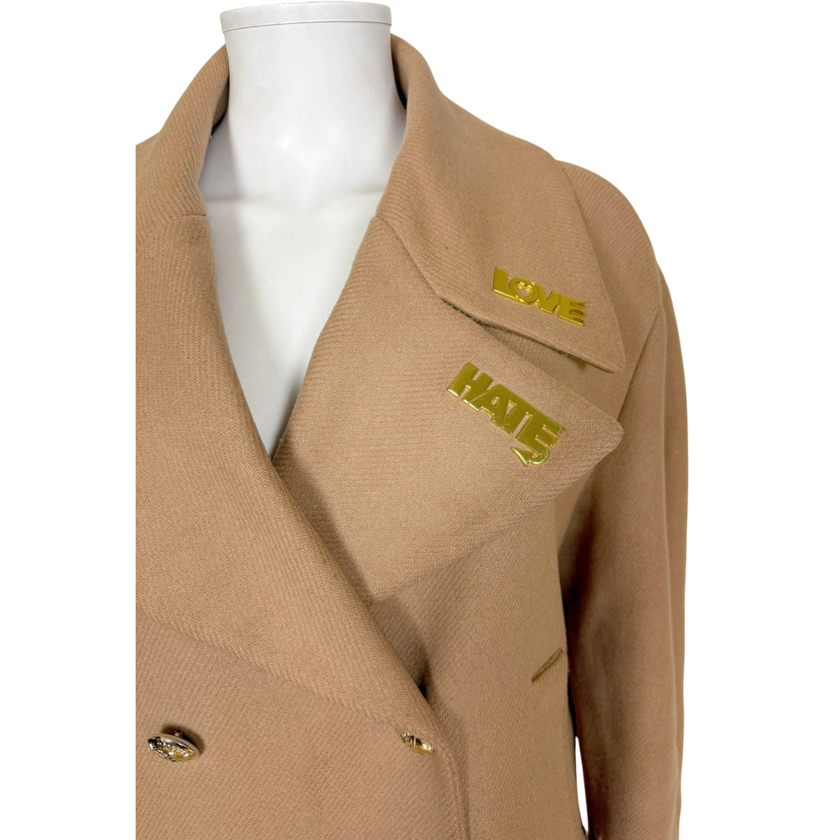 A close-up of two gold-tone lapel pins spelling 'LOVE' and 'HATE' on the collar, on a Love Moschino coat