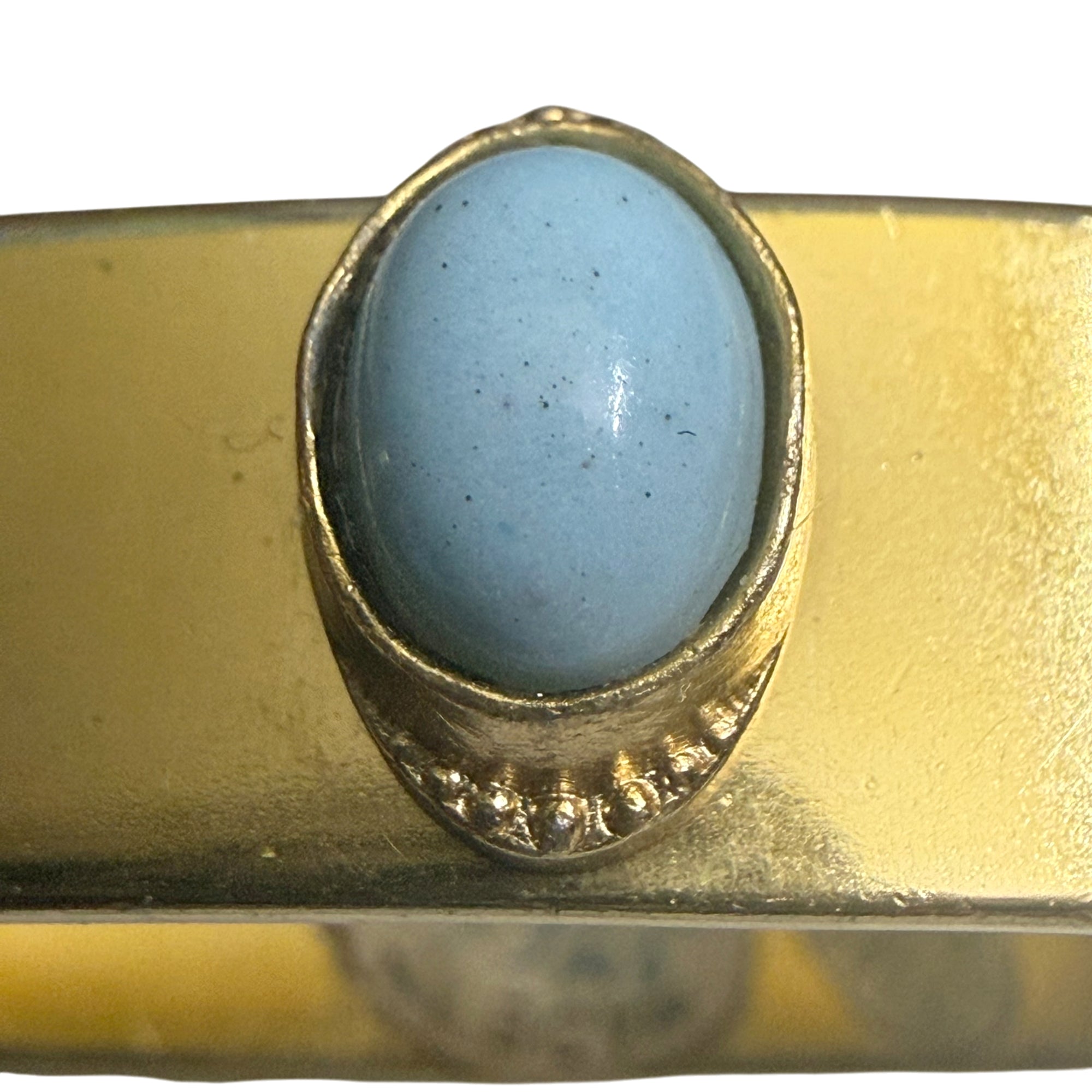 A close-up of a gold bangle bracelet featuring a large, oval-shaped light blue stone in a textured gold toned bezel setting.