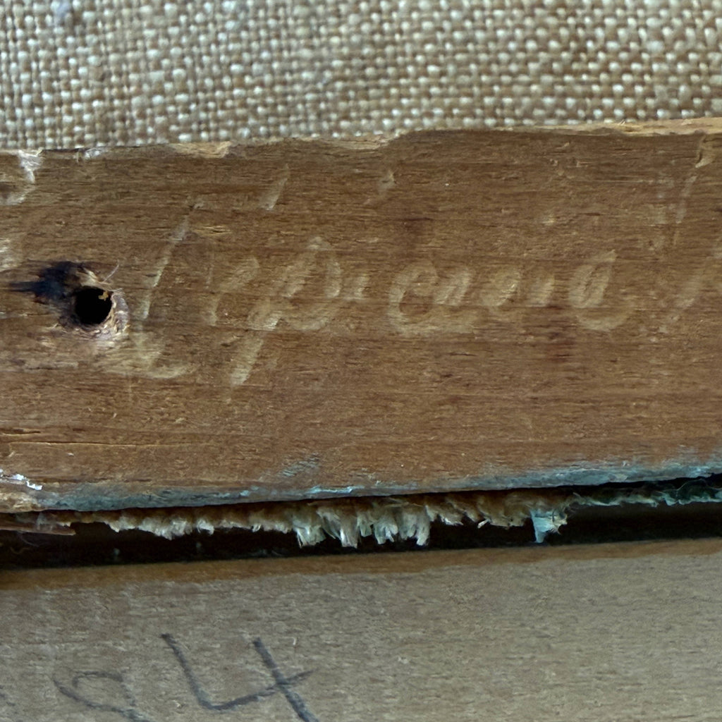 Close-up of the back corner of a wooden framed artwork, showing embossed script lettering on the wooden stretcher and canvas edge against a beige woven fabric background.